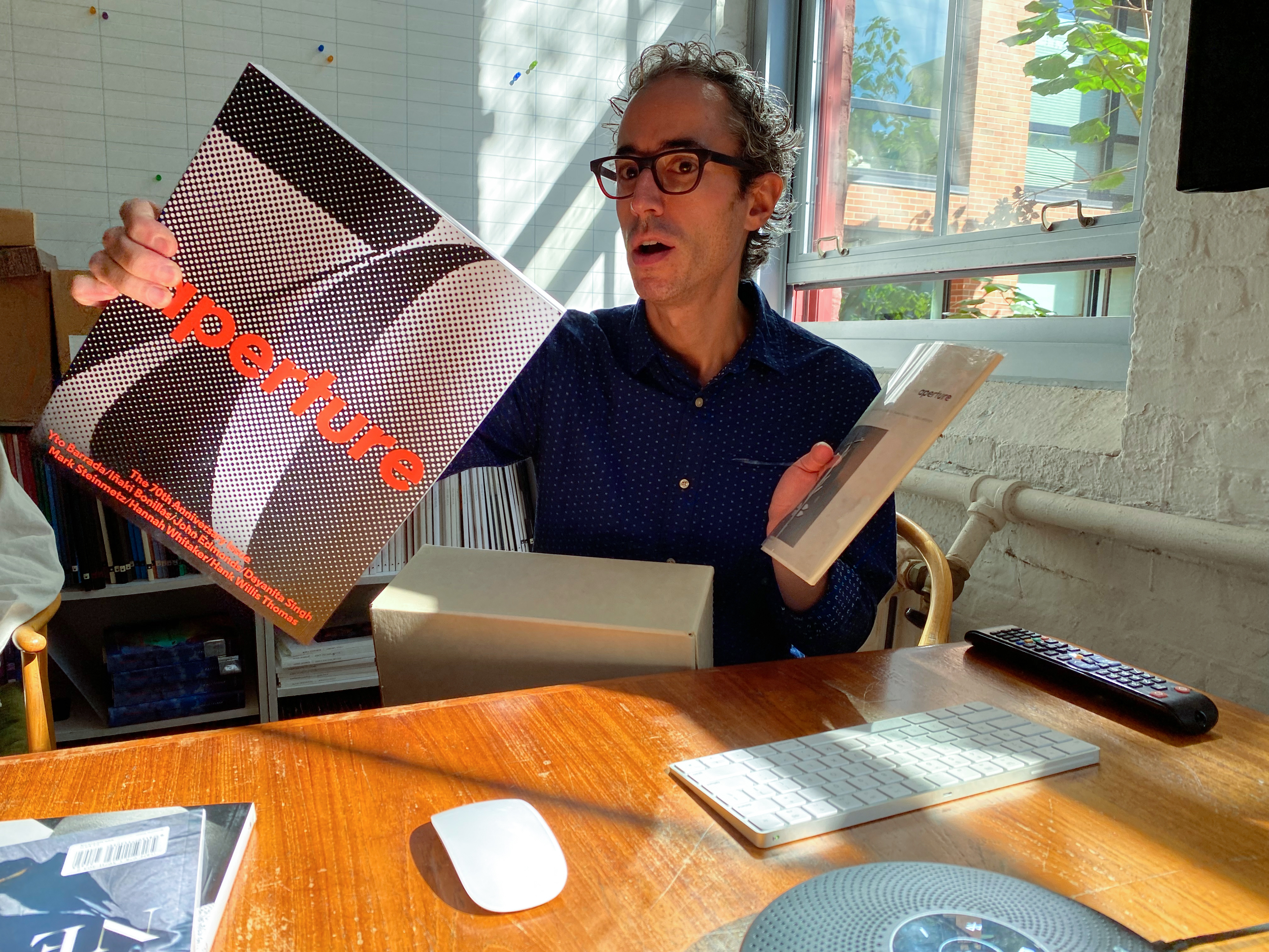 Brendan Embser,  Senior Managing Editor of Aperture, holds up multiple books while at a desk.