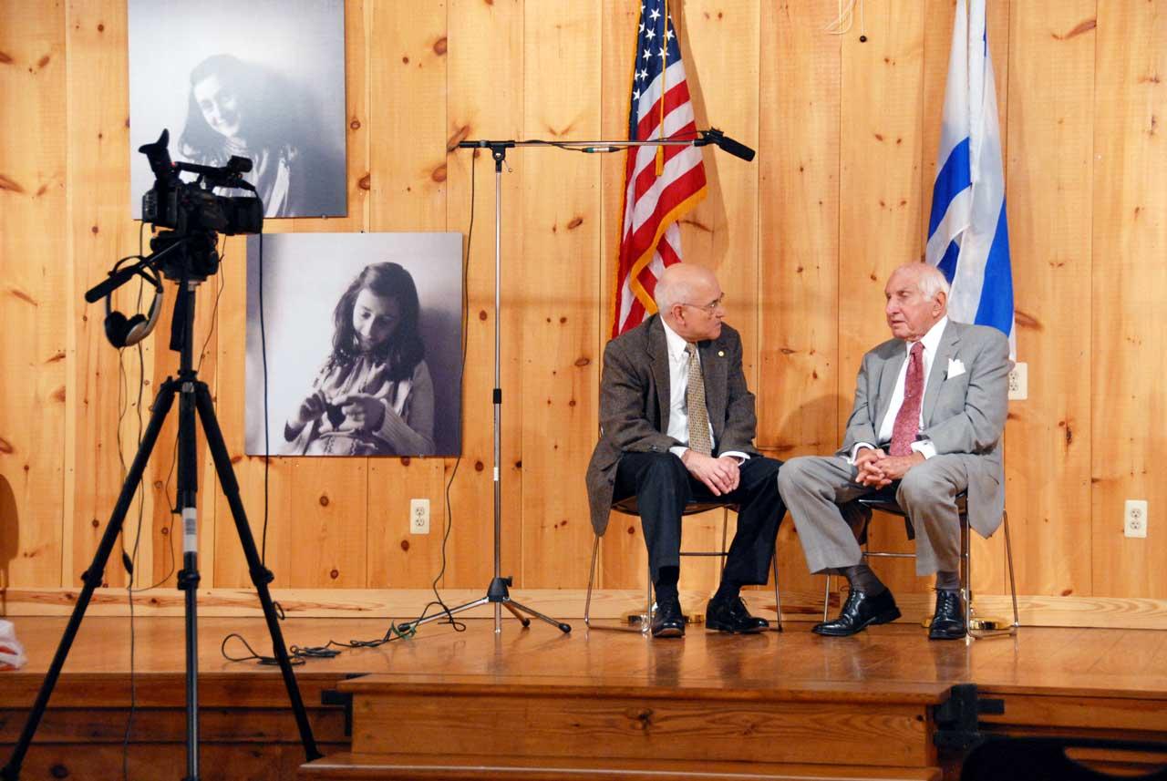Two men sit on a stage together with a camera pointed at them.