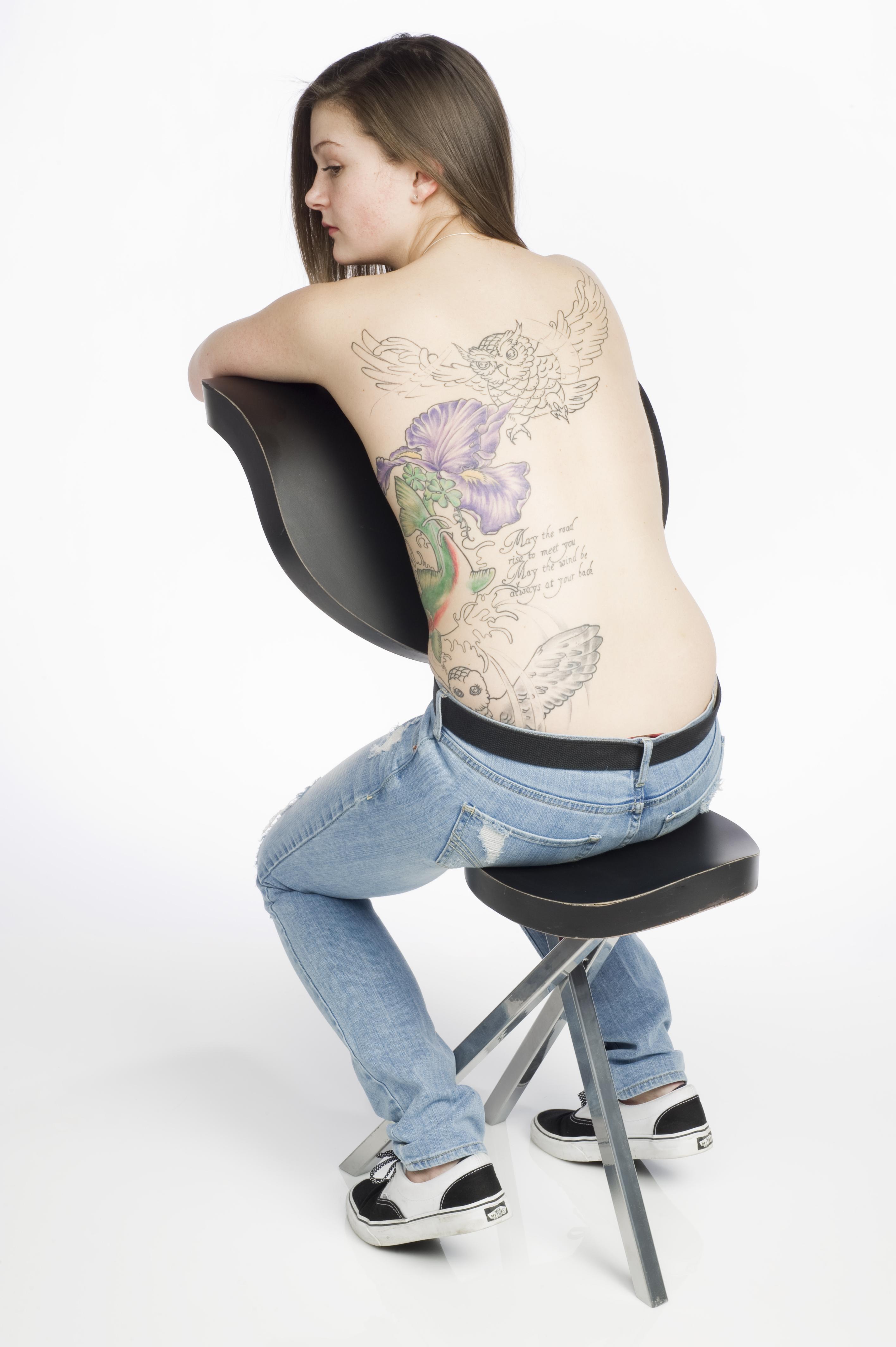 A shirtless person sits on a tattoo chair.