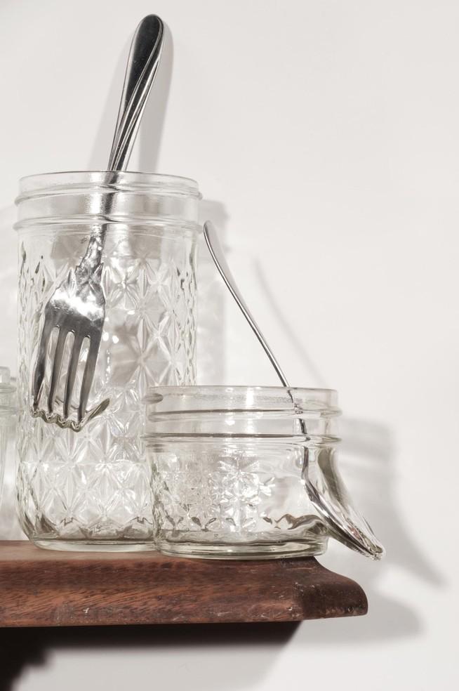 Glass jelly jars with silverware sits on a bracket mounted on a wall.